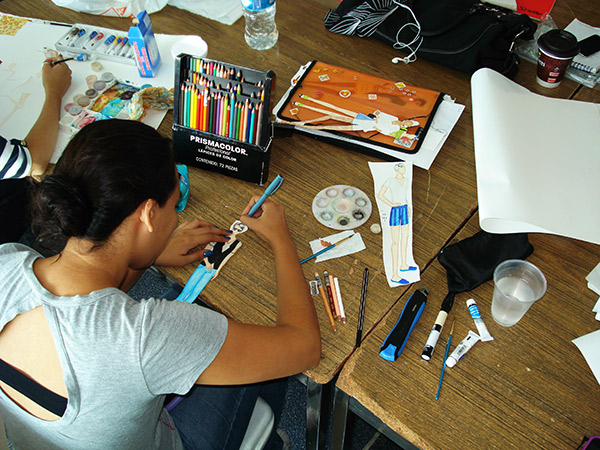 Creación de una colección para la clase de dibujo de figura humana usando la técnica de acuarela. Se observa una alumna dándole color a un figurín usando colores de lápiz de madera.