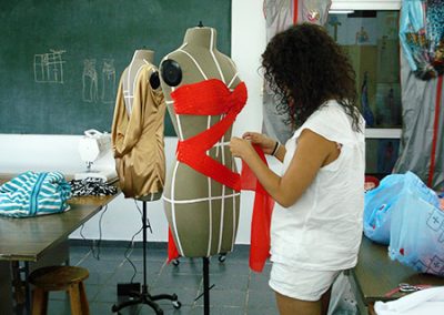 Creación de una prenda para la case de modelado sobre maniquí. La alumna está colocando una tela roja sobre el maniquí en forma de zigzag. En el salón se observan otros proyectos sobre los maniquies.