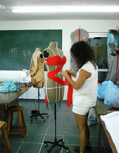 Creación de una prenda para la case de modelado sobre maniquí. La alumna está colocando una tela roja sobre el maniquí en forma de zigzag. En el salón se observan otros proyectos sobre los maniquies.