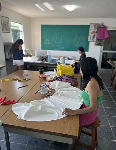 Elaboración de prendas para la clase de corte y confección. Se muestran 3 alumnas con sus telas sobre las mesas de corte.